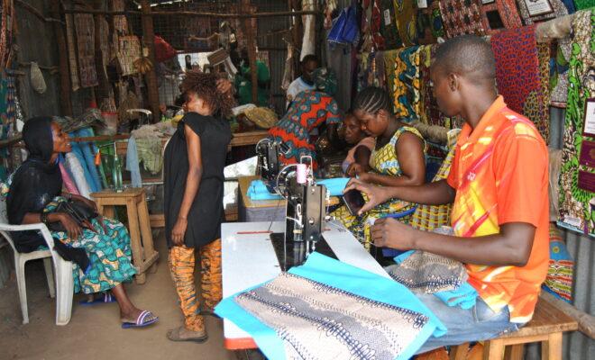 Biclere (standing) talks to a cliet at her shop while her employees work on other clothes.