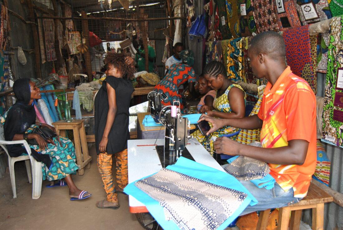 Biclere (standing) talks to a cliet at her shop while her employees work on other clothes.