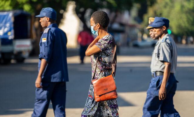 Chin'ono A woman wearing a Covid19 protective mask walks past a group of police officers on patrol in Bulawayo CBD, 1 April 2020. Credit photo : KB Mpofu / ILO