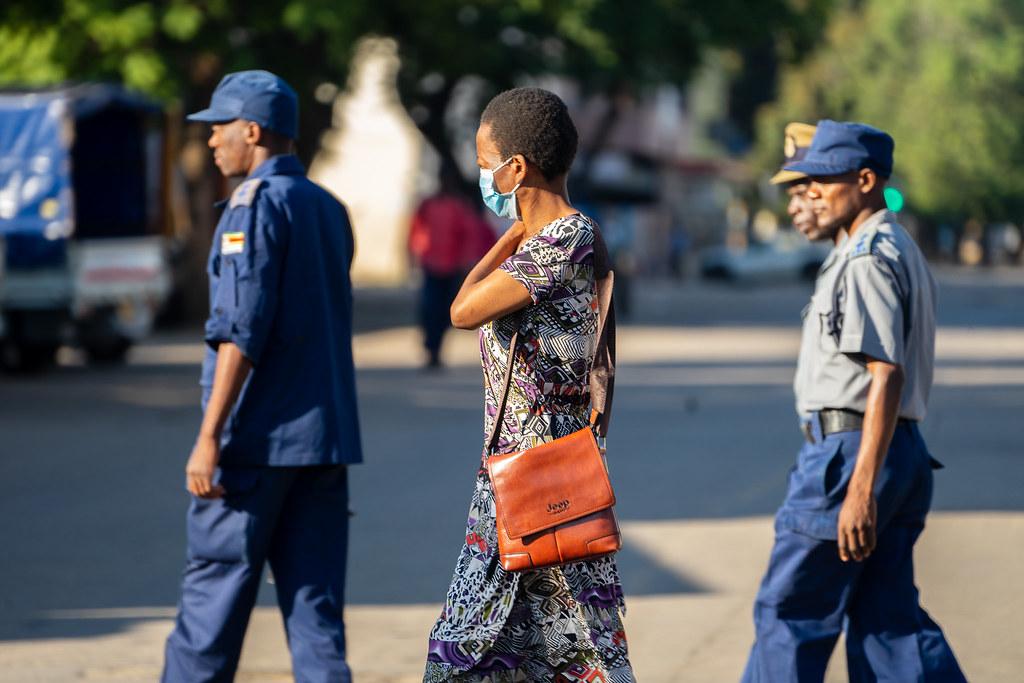 Chin'ono A woman wearing a Covid19 protective mask walks past a group of police officers on patrol in Bulawayo CBD, 1 April 2020. Credit photo : KB Mpofu / ILO
