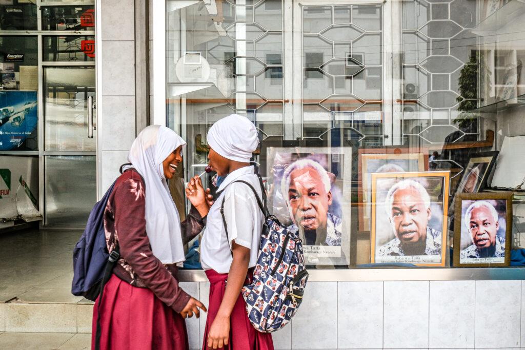 two school girls stand outside a shop in Arusha, Tanzania, with framed photos of Julius Nyerere displayed. Credit: Jaclynn Ashly.