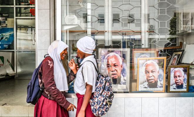 two school girls stand outside a shop in Arusha, Tanzania, with framed photos of Julius Nyerere displayed. Credit: Jaclynn Ashly.