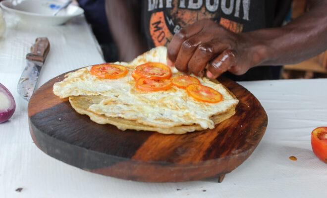 Fred Zziwa, a food vendor, adds tomatoes on top of fried eggs to make a rolex in Kabulengwa , Wakiso district, Uganda in April 2022. Zziwa says in the last three months prices of wheat flour and cooking oil have gone up he has to reduce the size of chapati to keep the same price for customers. Credit: Nakisanze Segawa/Global Press Journal.